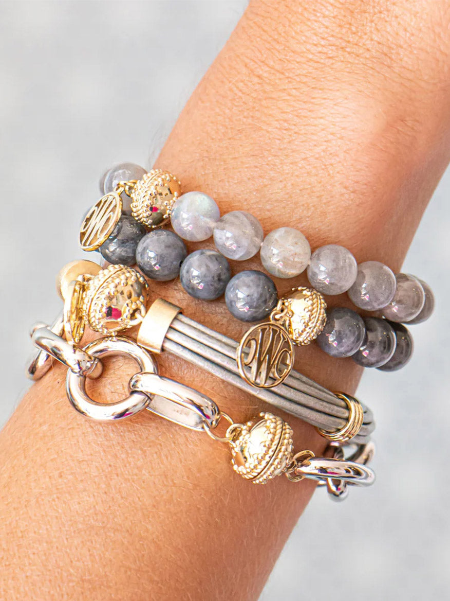 A wrist adorned with the Clara Williams Gold Coast Rhodium Bracelet and three gray gemstone beaded bracelets featuring 14K yellow gold accents, decorative charms, metal links in gold and silver, all finished with an elegant ClaraClasp.