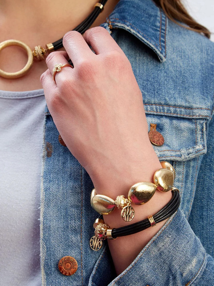 A person wearing a denim jacket, white shirt, 14K yellow gold ring, a gold necklace with a round pendant, and the Clara Williams Gold Rush Large Teardrop Stretch Bracelet.