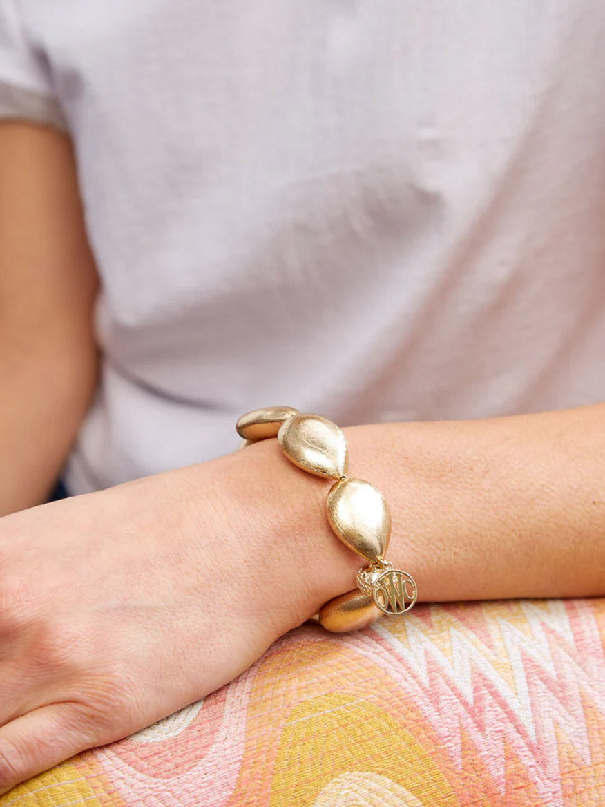 A person in a white shirt rests their hand on a patterned surface, showcasing the Clara Williams Gold Rush Large Teardrop Stretch Bracelet.