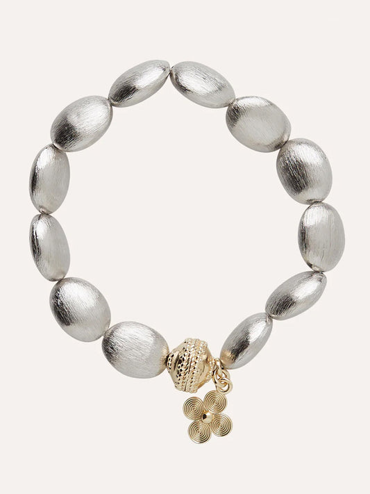 The Clara Williams Gold Rush Rhodium Stretch Bracelet features oval silver beads, brushed rhodium beads, a gold-toned textured charm, and a small spiral pendant.