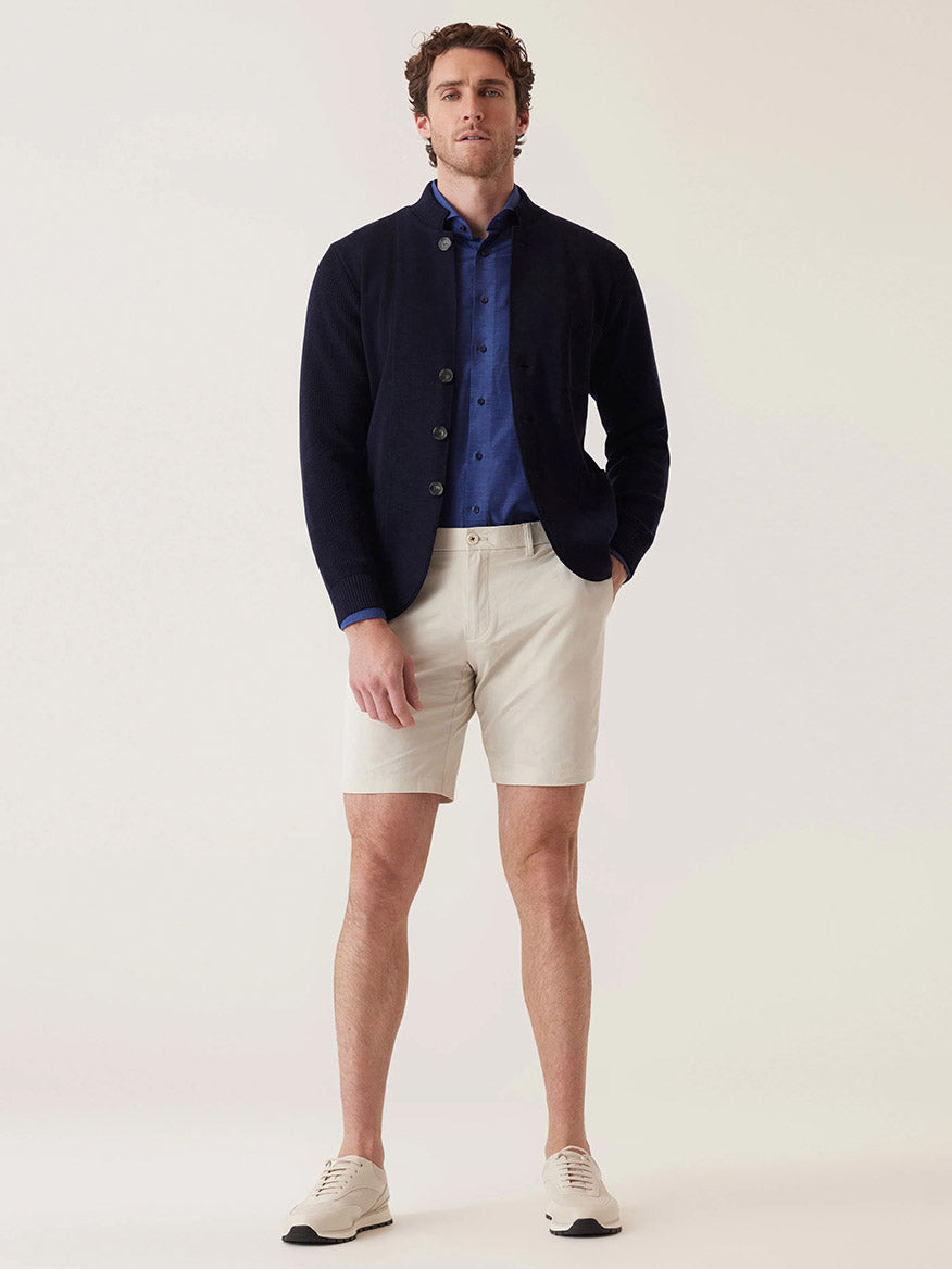 A man exudes performance-driven sophistication in his navy blazer, blue shirt, DEKE Galaxy Bermuda Ultra 4X Stretch Twill shorts in Silver, and light-colored sneakers, showcasing a tailored fit that melds comfort and style against a plain background.