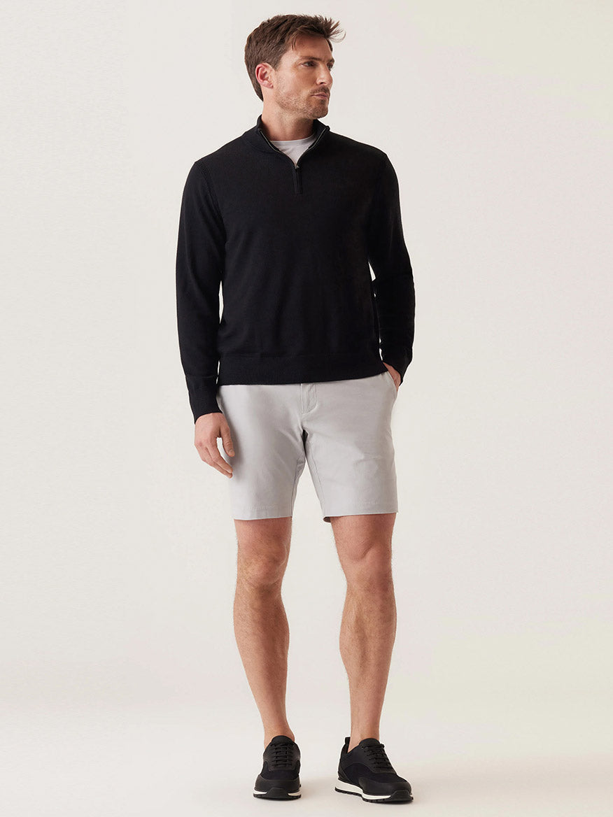 A man stands against a plain background wearing a black quarter-zip sweater, black sneakers, and DEKE Galaxy Bermuda Ultra 4X Stretch Twill shorts in Stone with a tailored fit.