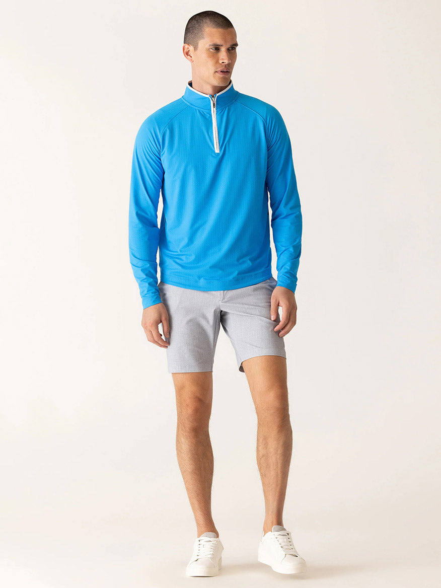A man wears a bright blue quarter-zip long sleeve shirt and DEKE Galaxy Bermuda Nano Nylon Warp Knit shorts in Silver, paired with white sneakers, standing against a plain light background.