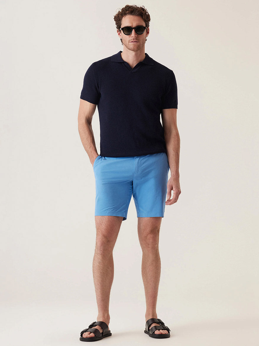 A man wearing sunglasses, a short-sleeve navy polo, DEKE Galaxy Bermuda Ultra 4X Stretch Twill shorts in Lake Blue with a tailored fit, and black sandals stands with hands in pockets against a plain light background.