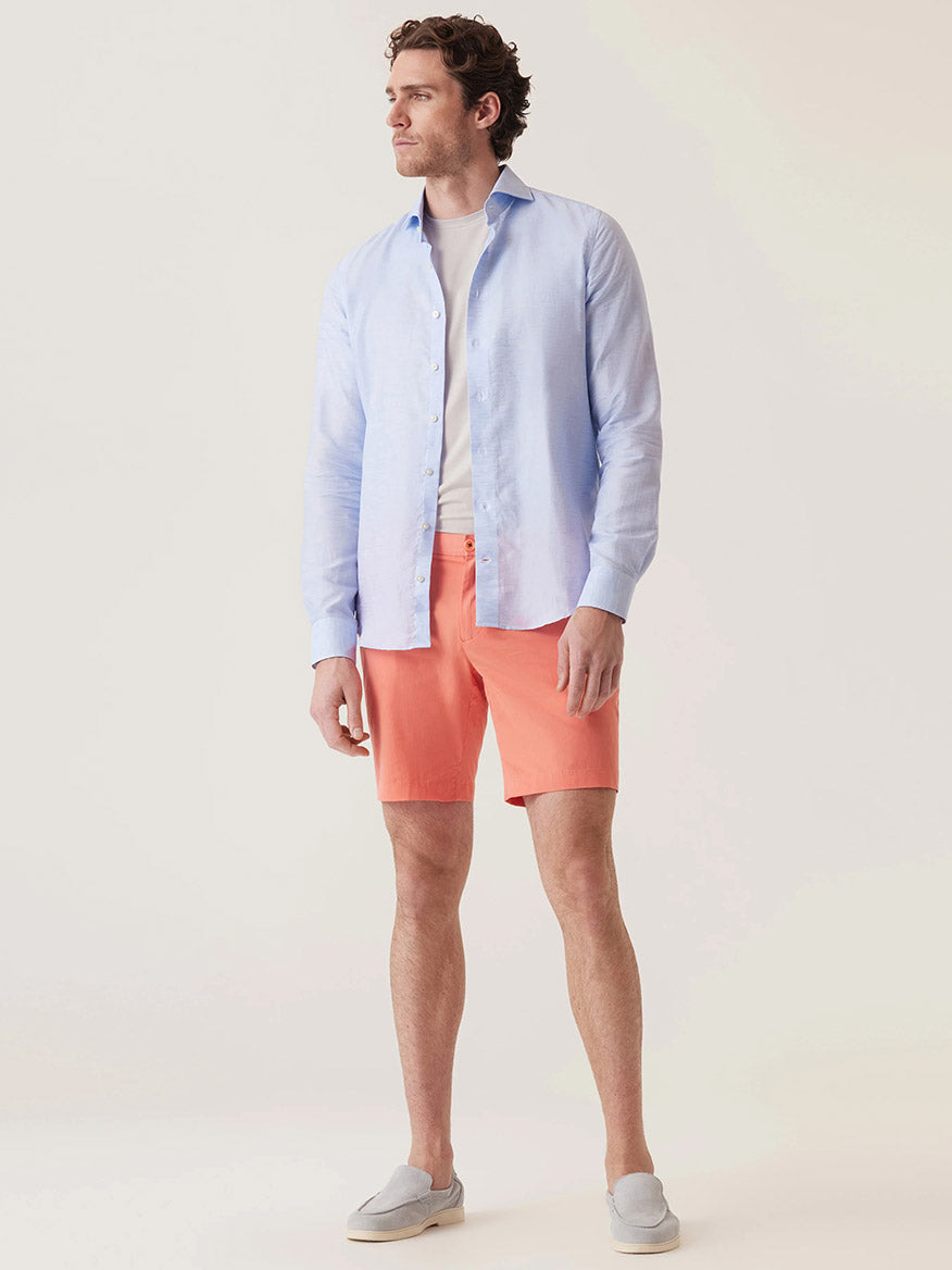 A man stands against a neutral background wearing a light blue button-up shirt over a white t-shirt, gray slip-on shoes, and the DEKE Galaxy Bermuda Ultra 4X Stretch Twill shorts in Sunset with a tailored fit.