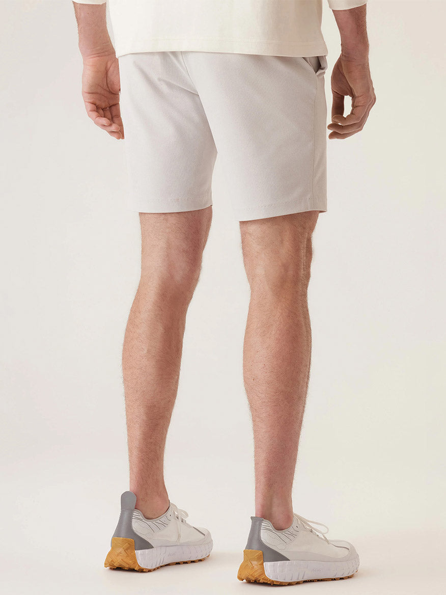 A person is seen from behind wearing DEKE Momentum Mobile Bermuda Pulse Pique Warp Knit shorts in Frost Grey, teamed with a beige shirt and white sneakers with gray accents and tan soles, against a plain background.