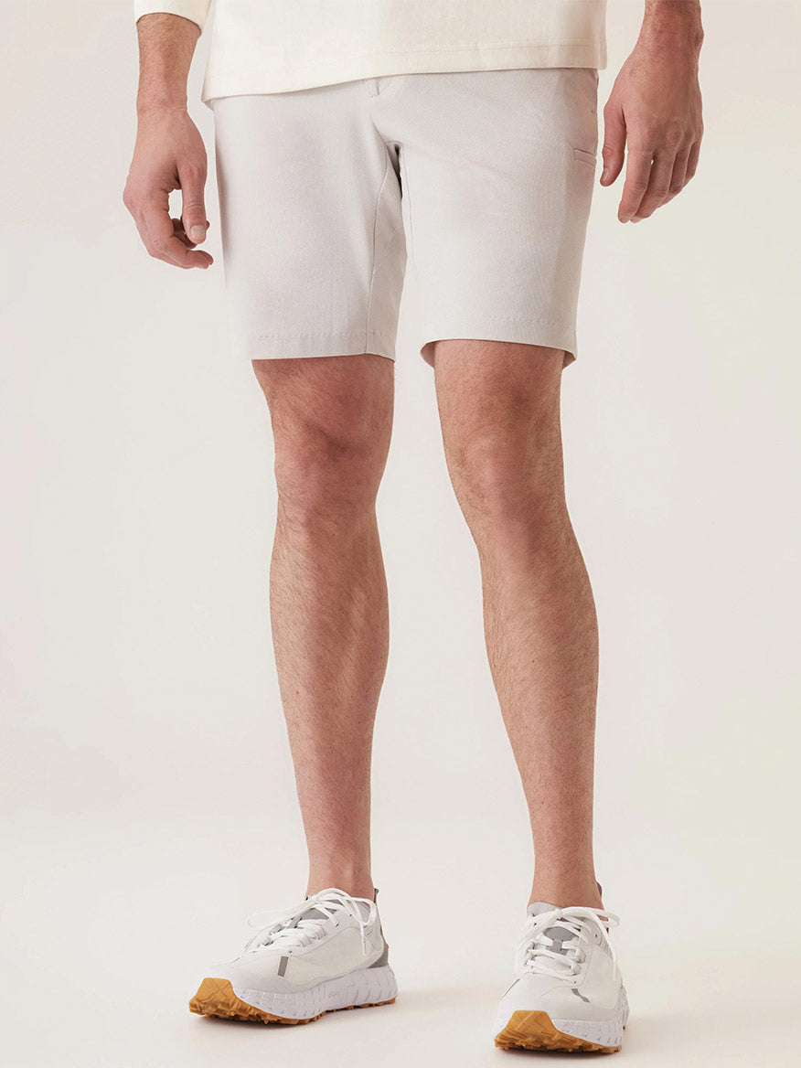 A person showcases a tailored outfit featuring a white long-sleeve shirt, DEKE Momentum Mobile Bermuda Pulse Pique Warp Knit shorts in Frost Grey, and white sneakers with brown soles, set against a neutral background.