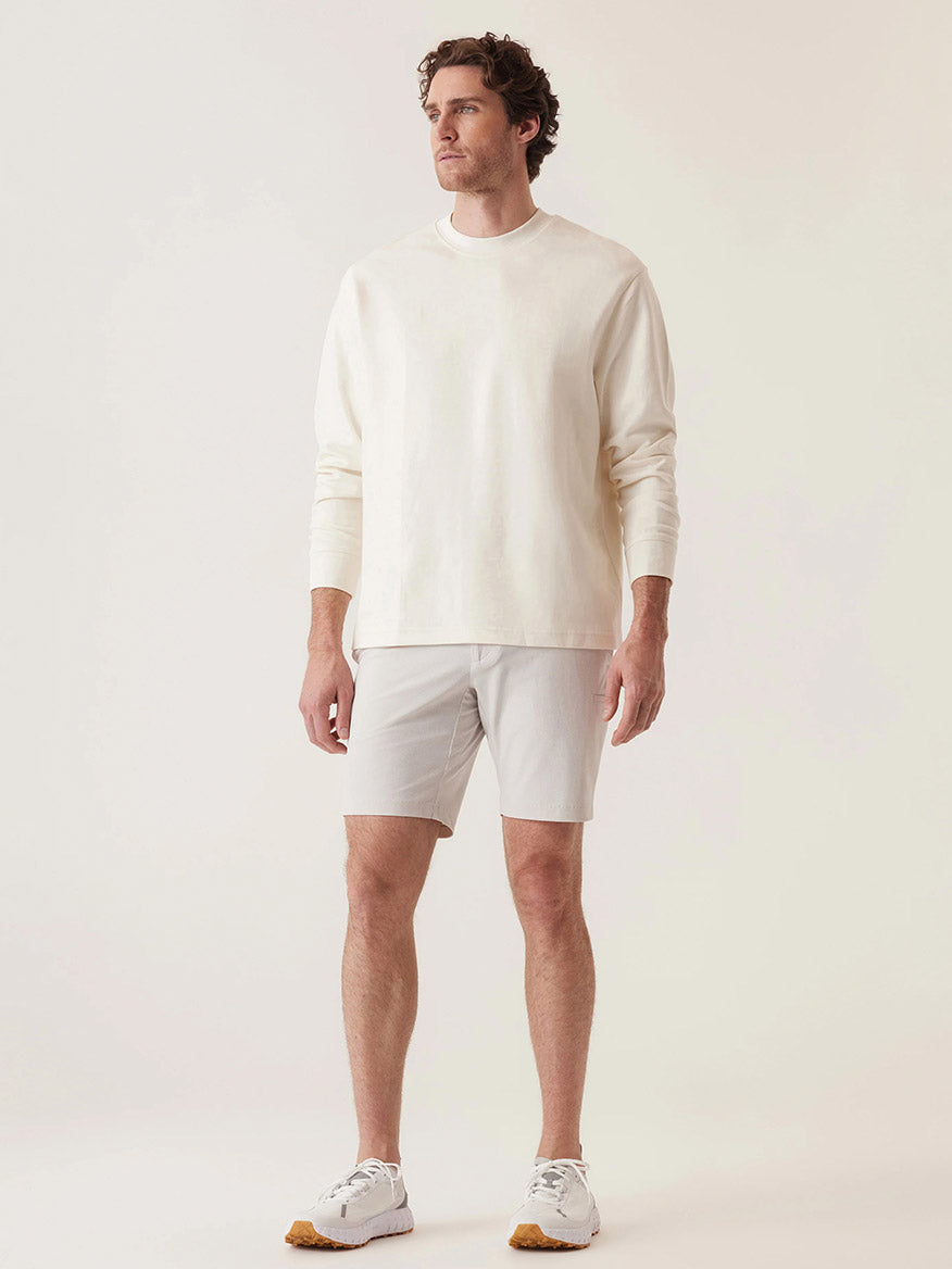 A man exudes casual elegance in a tailored fit cream long-sleeve shirt, paired with DEKE's Momentum Mobile Bermuda Pulse Pique Warp Knit shorts in Frost Grey and white sneakers against a neutral backdrop.