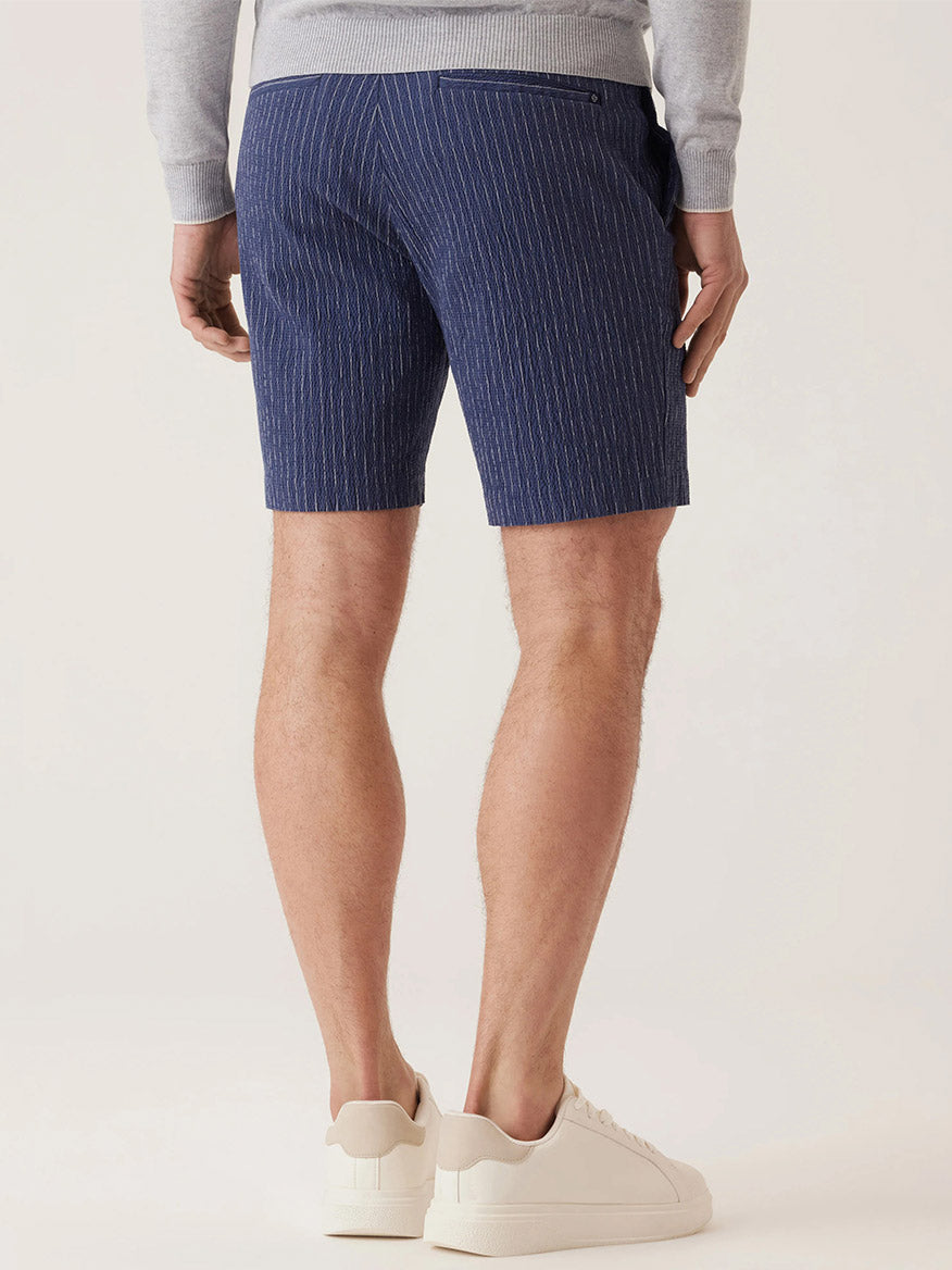 Rear view of a person in DEKE Oasis Bermuda Supernatural Seersucker shorts (Indigo Heather Pinstripe), gray long-sleeve top, and white sneakers against a plain background; this warm-weather essential exudes effortless style.