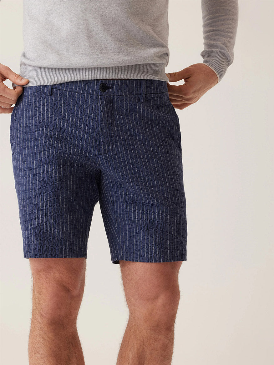 Clad in the DEKE Oasis Bermuda Supernatural Seersucker in Indigo Heather Pinstripe, a person embodies warm-weather style against a neutral background, confidently resting their hands on their hips.