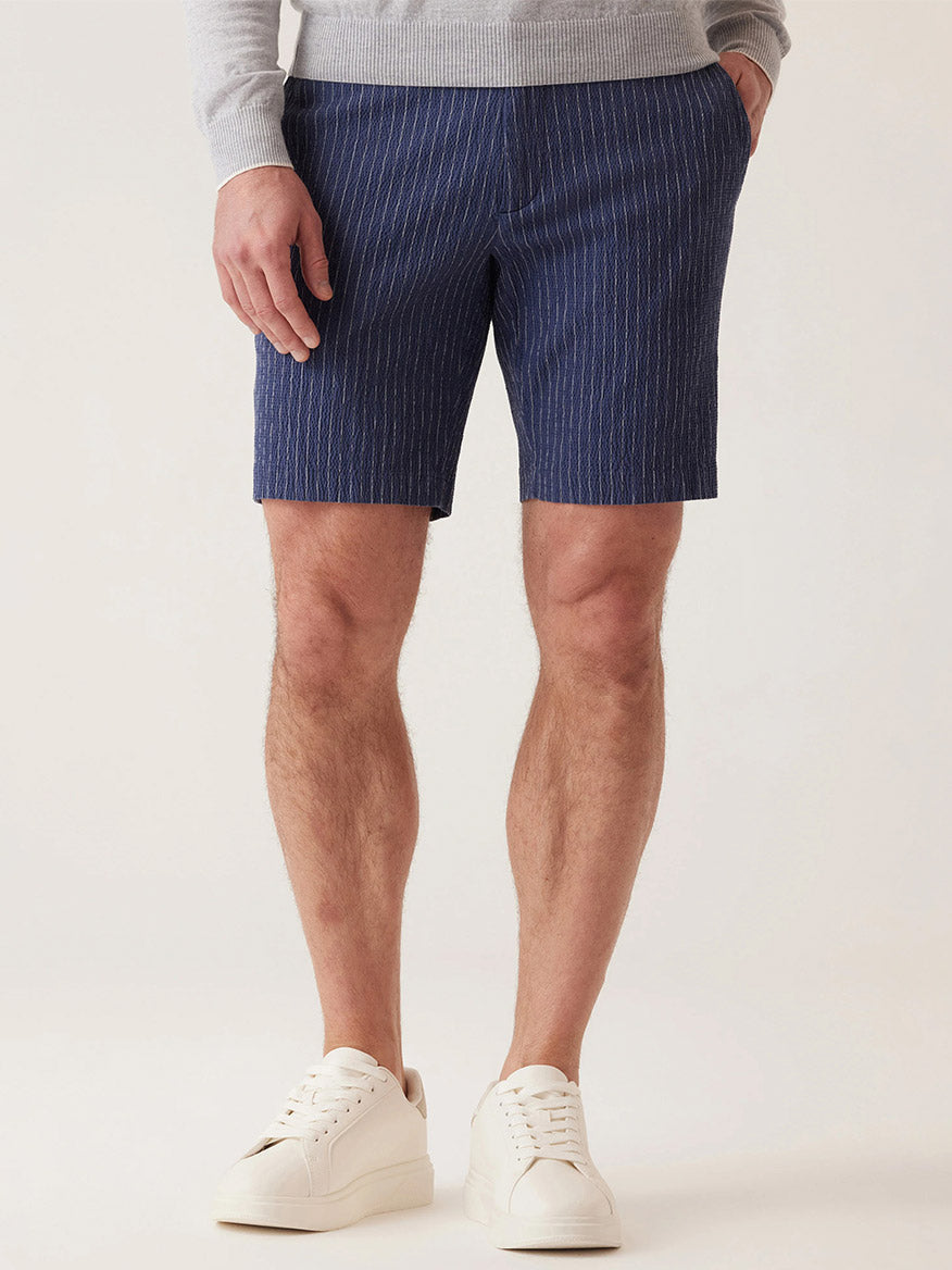 A person effortlessly showcases the DEKE Oasis Bermuda Supernatural Seersucker in Indigo Heather Pinstripe, paired with a gray sweater and white sneakers, standing against a plain background.