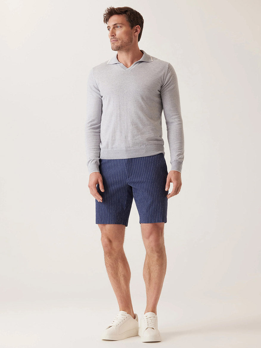 A man in a light gray sweater, blue shorts, and white sneakers stands against a plain background, effortlessly embodying the warm-weather style of DEKE Oasis Bermuda Supernatural Seersucker in Indigo Heather Pinstripe as he faces slightly to the left.