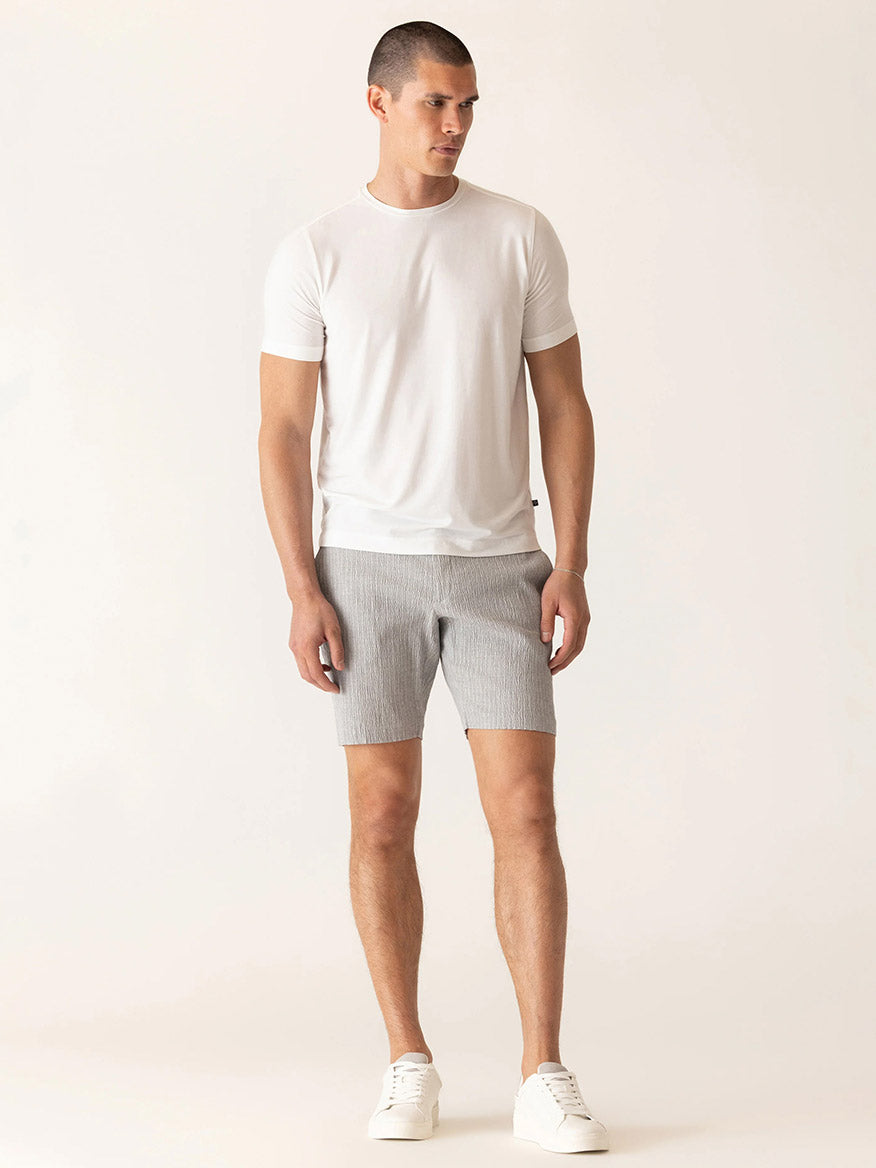 A man stands in a neutral pose wearing a white t-shirt, white sneakers, and DEKE Oasis Bermuda Supernatural Seersucker shorts in Grey/Natural against a plain light background.