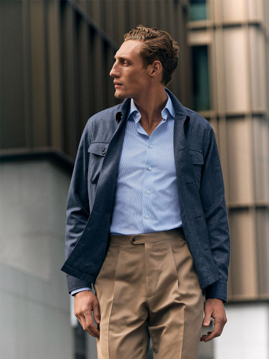 A man in an Eton Contemporary Fit Fine Striped Signature Twill Dress Shirt in Light Blue, dark jacket, and beige pleated trousers stands outside a modern building, embodying a business classic look as he gazes to his left.