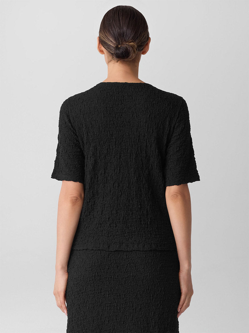 A person stands facing away from the camera in the Eileen Fisher Bubble Jacquard Crew Neck Top in Black, with their hair neatly tied in a low bun.
