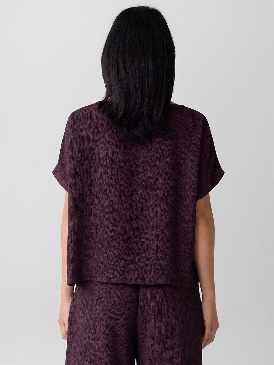 A person with shoulder-length dark hair stands with their back to the camera, wearing the Eileen Fisher Woven Plissé Dolman Sleeve Top in Blackberry and matching pants.