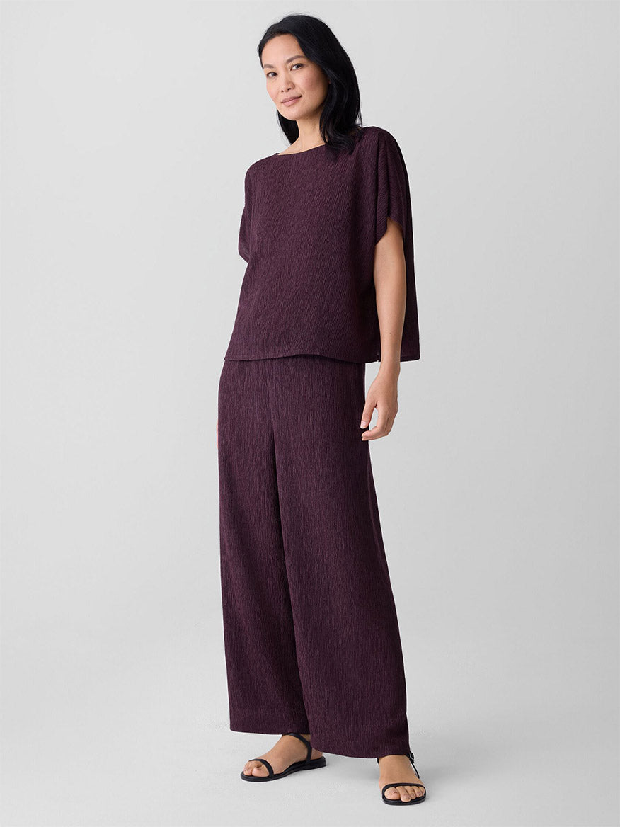 A woman wears the Eileen Fisher Woven Plissé Dolman Sleeve Top in Blackberry with matching wide-leg pants and black sandals, standing against a light background.