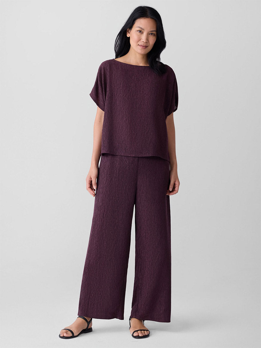 A woman stands against a plain background wearing black sandals and the Eileen Fisher Woven Plissé Wide-Leg Pant in Blackberry, paired with a loose-fitting, textured dark purple top.