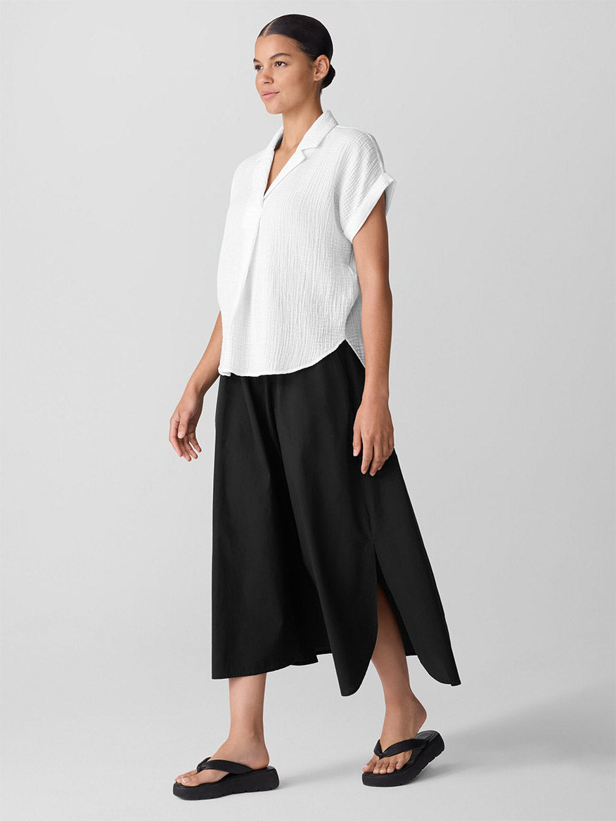 A woman stands sideways wearing an Eileen Fisher Organic Cotton Lofty Gauze Notch Collar Top in White, a black midi skirt with side slits, and black sandals against a plain background.
