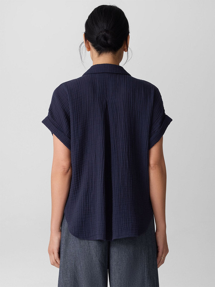 A person with dark hair in a low bun is shown from behind wearing the Eileen Fisher Organic Cotton Lofty Gauze Notch Collar Top in Ink and gray pants, standing against a plain light background.