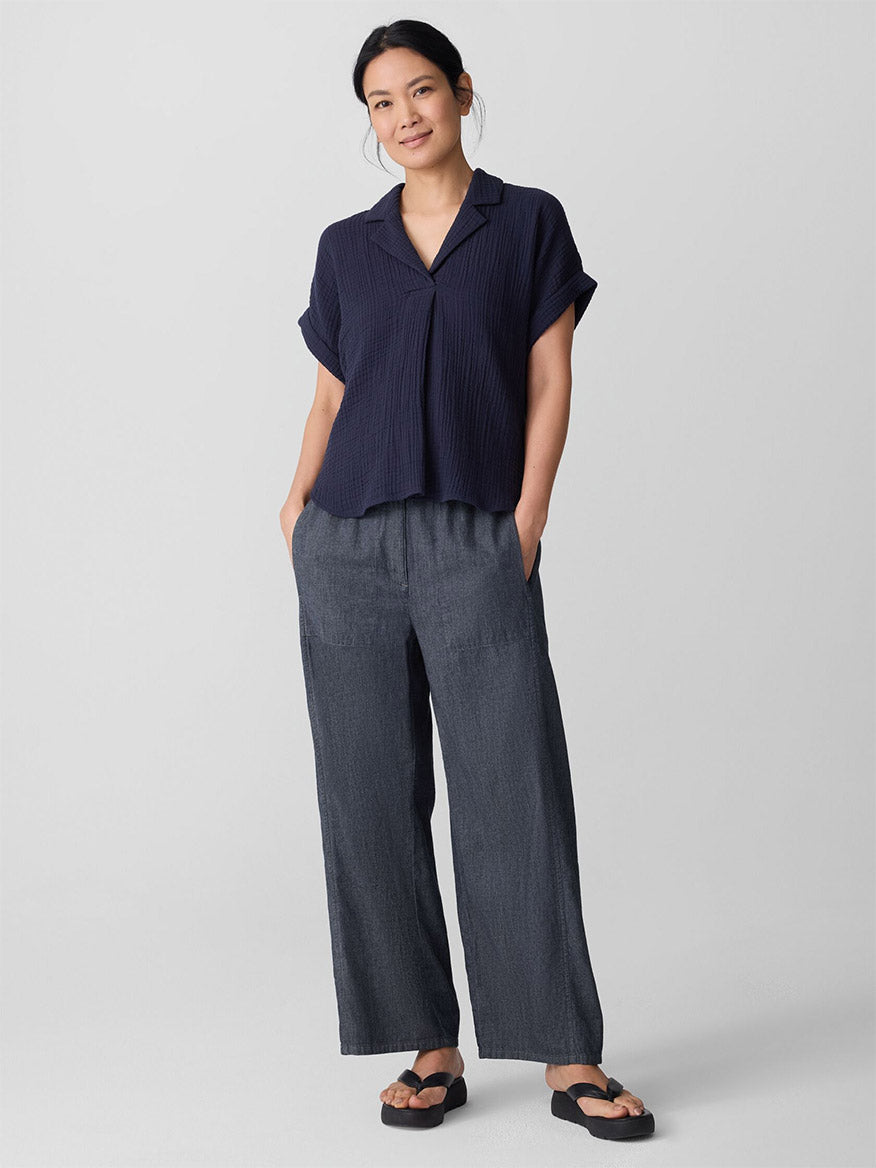 A woman stands against a plain background wearing the Eileen Fisher Organic Cotton Lofty Gauze Notch Collar Top in Ink, styled with loose gray pants and black sandals, her hands casually placed in her pockets.