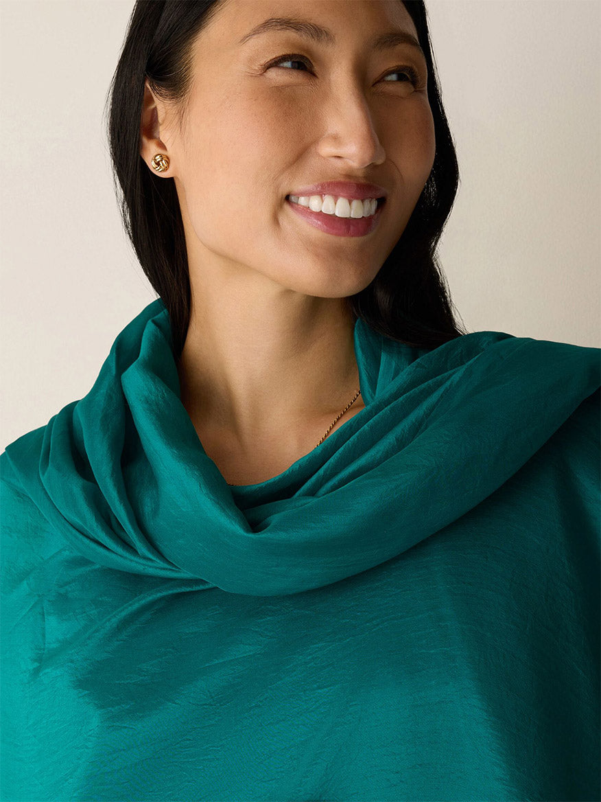 A woman with long dark hair smiles while wearing the Eileen Fisher Washed Organic Silk Scarf in Teal and gold stud earrings, set against a plain light background.
