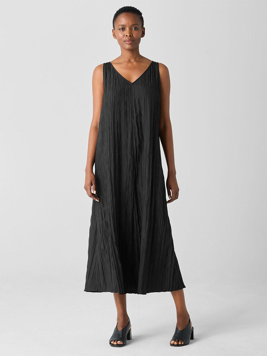 A person stands in an Eileen Fisher Crushed Silk V-Neck Dress in black, paired with black open-toe shoes, against a plain background.