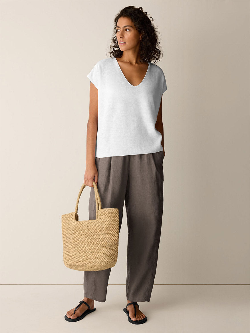 A woman stands indoors wearing the Eileen Fisher Organic Linen Cotton V-Neck Top in white, paired with loose brown pants, black sandals, and holding a woven straw bag.