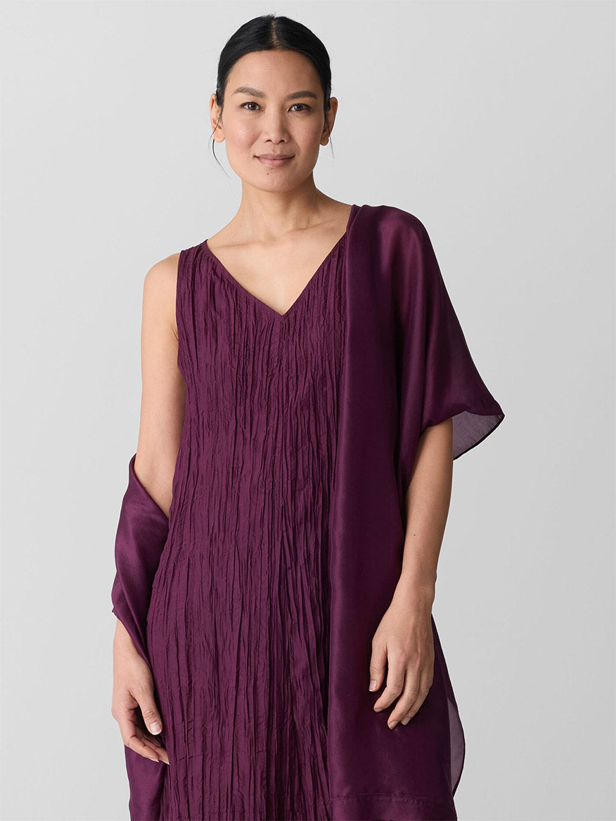 A woman stands against a light gray background, elegantly draping the Eileen Fisher Washed Transitional Silk Scarf in Blackberry over one shoulder while wearing a sleeveless, deep purple textured dress.