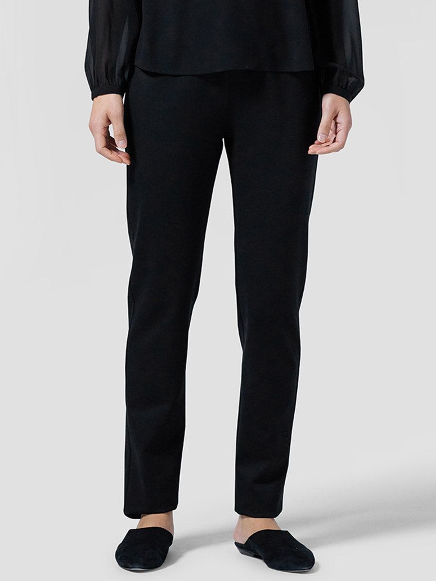A person stands against a plain background, dressed in all black, wearing Eileen Fisher Slim Ankle Pants with a yoke design in stretch knit fabric featuring an elastic waist, paired with a matching top and shoes.