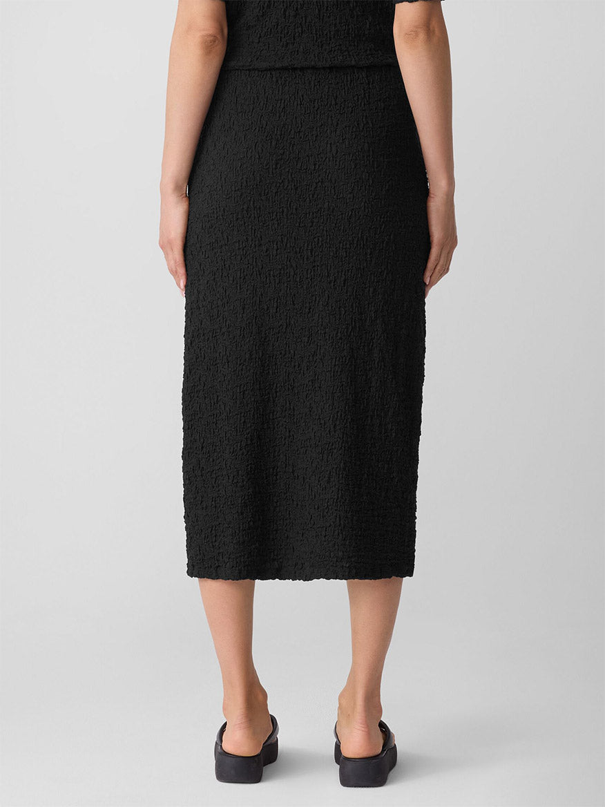 A person faces away, wearing the Eileen Fisher Bubble Jacquard Pencil Skirt in Black with black shoes against a plain background.