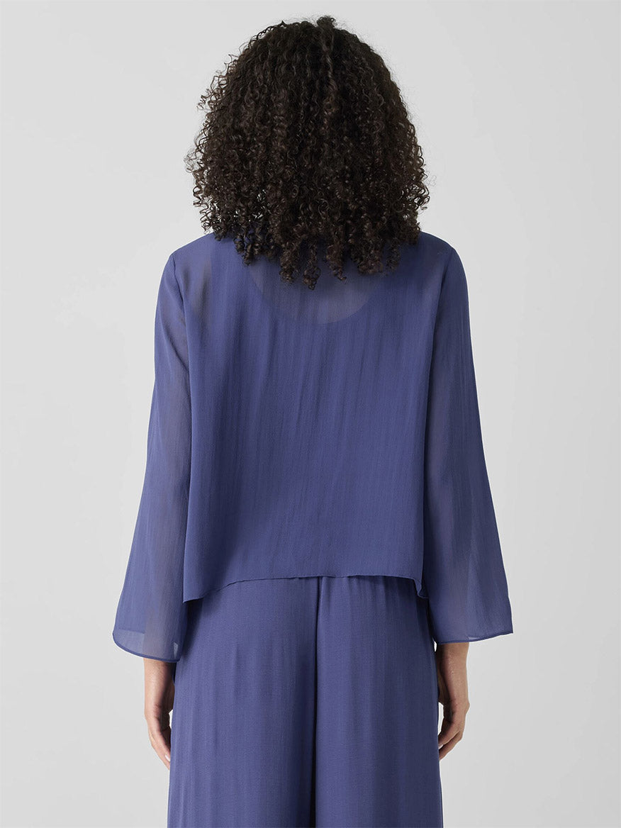 A person with curly hair wears an Eileen Fisher Sheer Silk Georgette Tie Jacket in Bluette and matching pants. Seen from behind against a plain background, delicate cascading ties add an elegant touch to the ensemble.