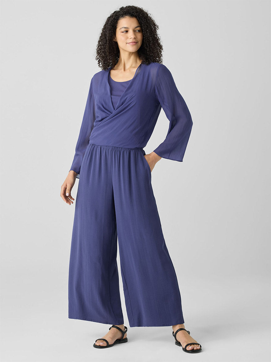 Dressed in the Eileen Fisher Sheer Silk Georgette Tie Jacket in Bluette, the individual poses with one hand in a pocket, showcasing style with its long sleeves and wide-legged pants. This piece adds an elegant touch and is perfect for any chic wardrobe.