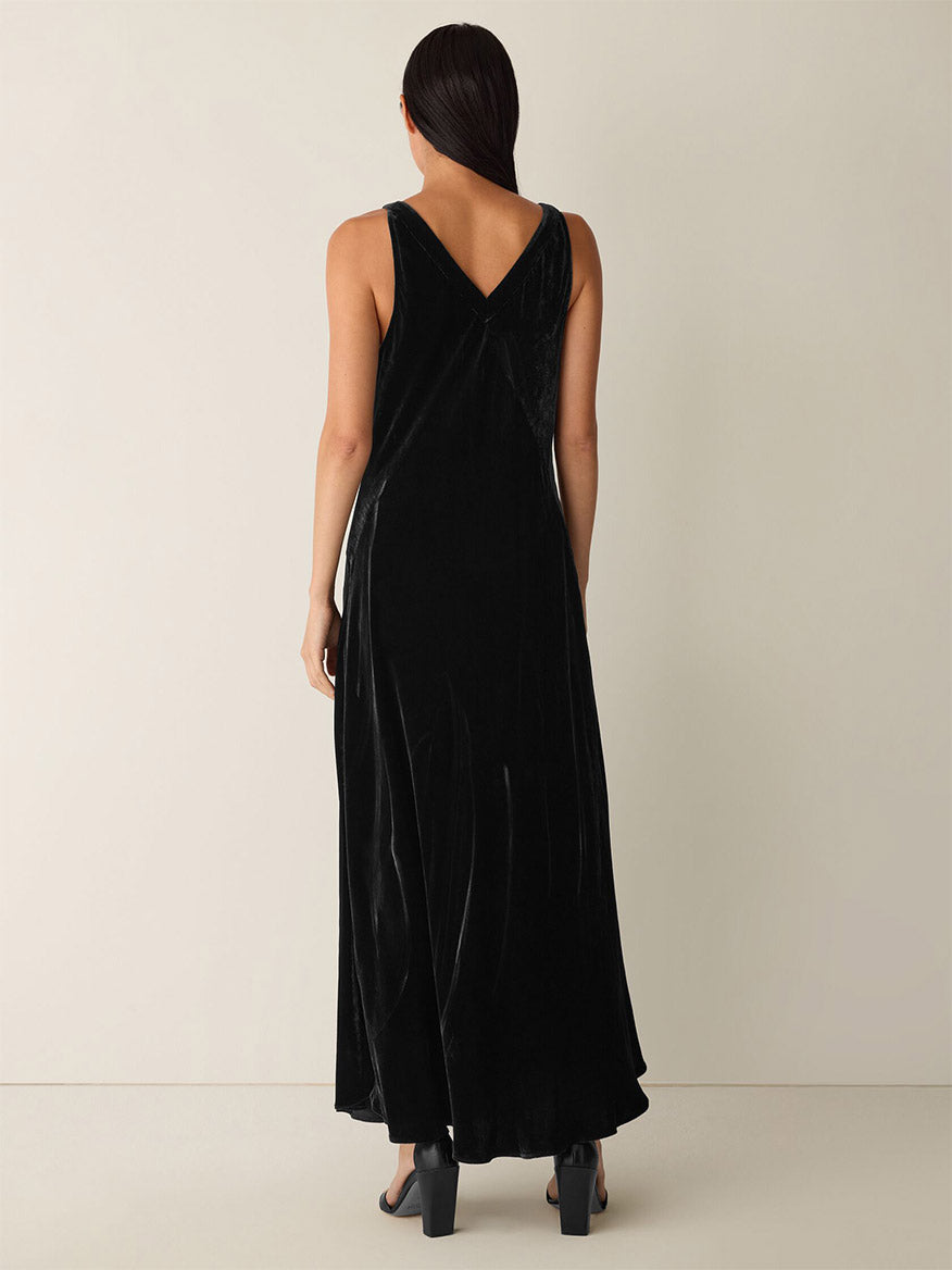 A person with long dark hair stands with their back to the camera against a plain light background, wearing the Eileen Fisher Velvet V-Neck Dress in Black and black heels.