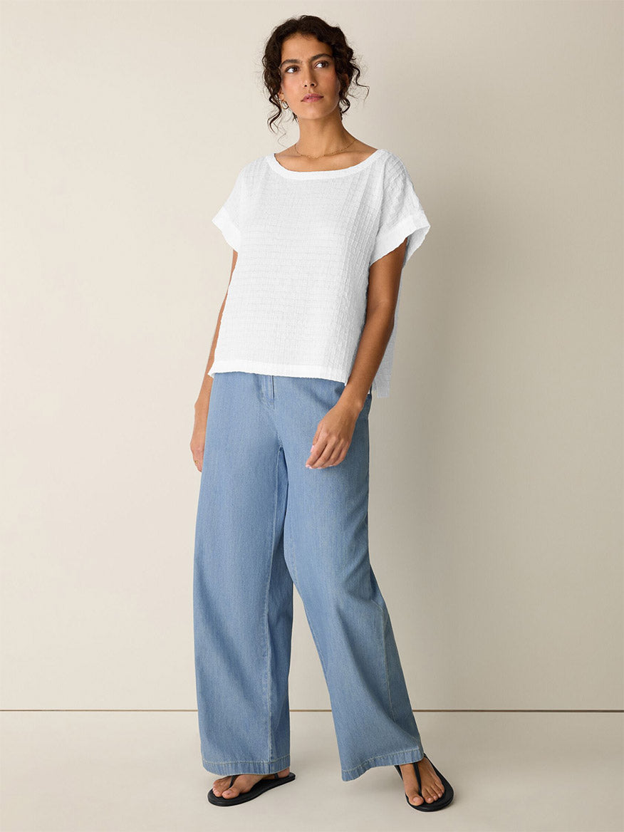 A woman stands against a plain wall wearing the Eileen Fisher Organic Cotton Voile Box Square Top in White, light blue wide-leg jeans, and black sandals.