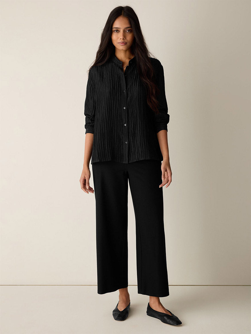 A woman stands against a plain light wall, wearing a black button-up pleated shirt, black flats, and Eileen Fisher Washable Stretch Crepe Wide-Leg Pant in Black with an elastic waist.