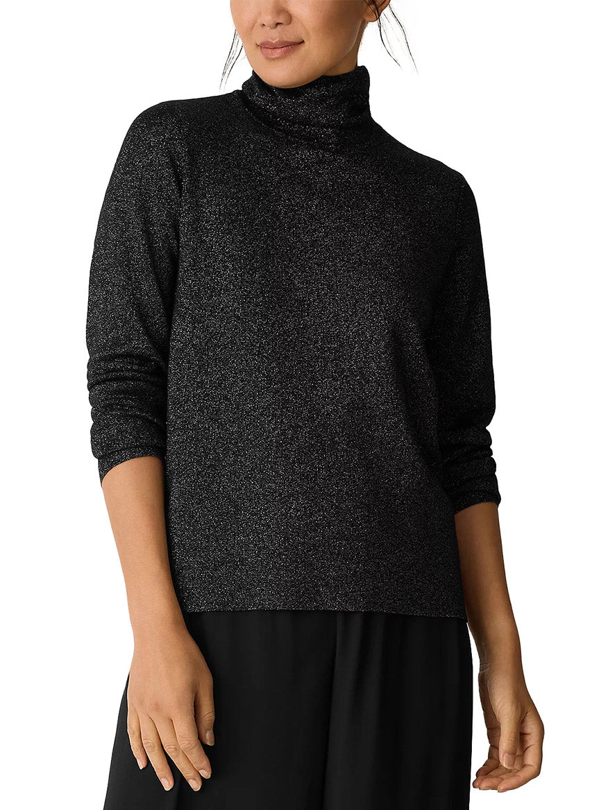 A person wearing the Eileen Fisher Wool Lurex Turtleneck Sweater in Black with black pants, standing against a white background.