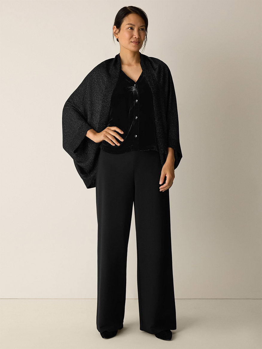 A woman stands against a plain background wearing the Eileen Fisher Wool Shimmer Serape in Black over a black blouse and wide-leg black pants.
