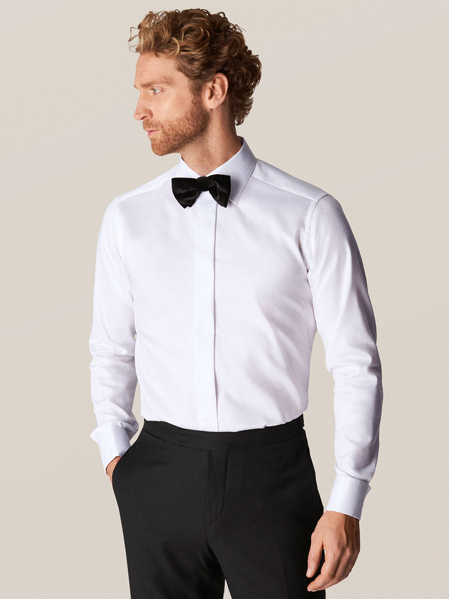 A man in an Eton White Dobby Formal Shirt with a black bow tie and black trousers stands against a plain background, looking to his left.
