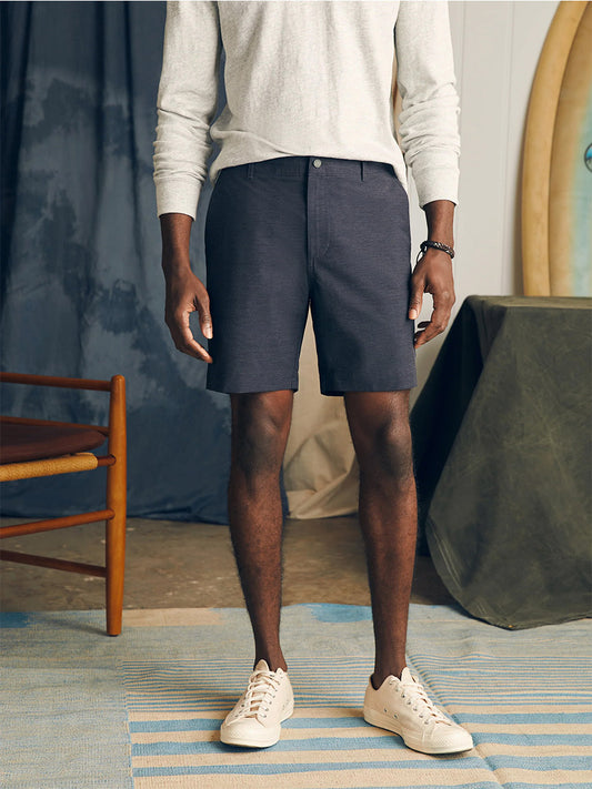 A man standing in a casual pose wearing a light grey sweatshirt, Faherty Brand All Day Shorts in Charcoal made of eco-friendly fabric, and white sneakers.