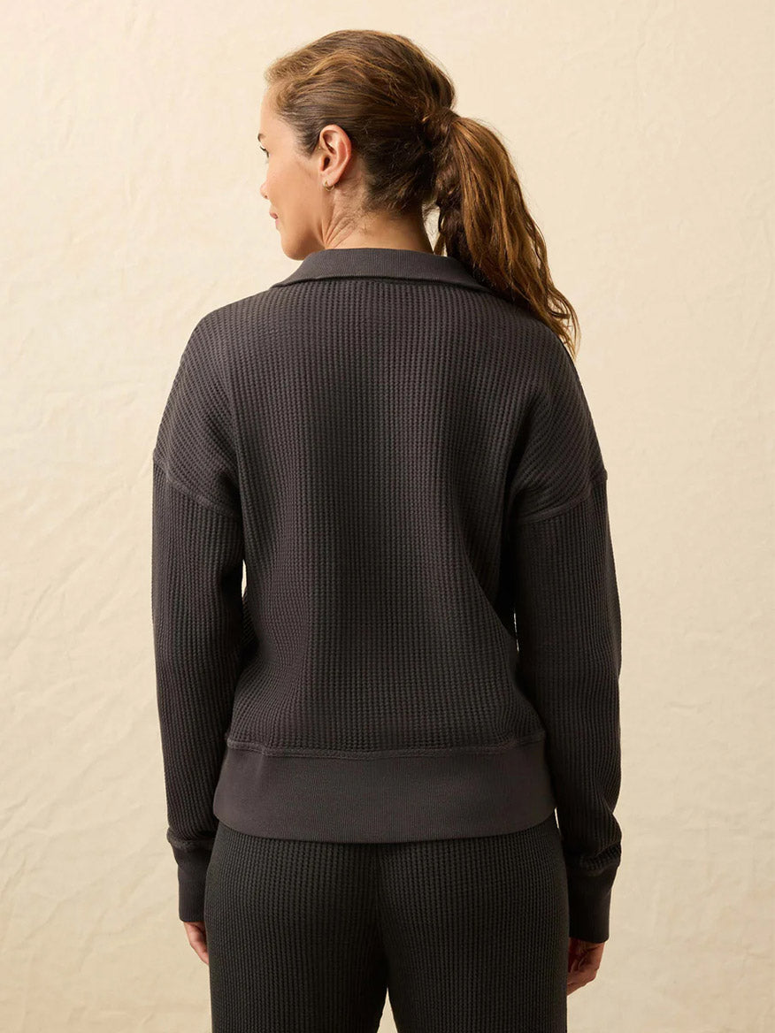 A woman with light brown hair in a ponytail is seen from the back wearing the Faherty Brand Après Waffle Henley in Washed Black, paired with matching pants against a neutral background.