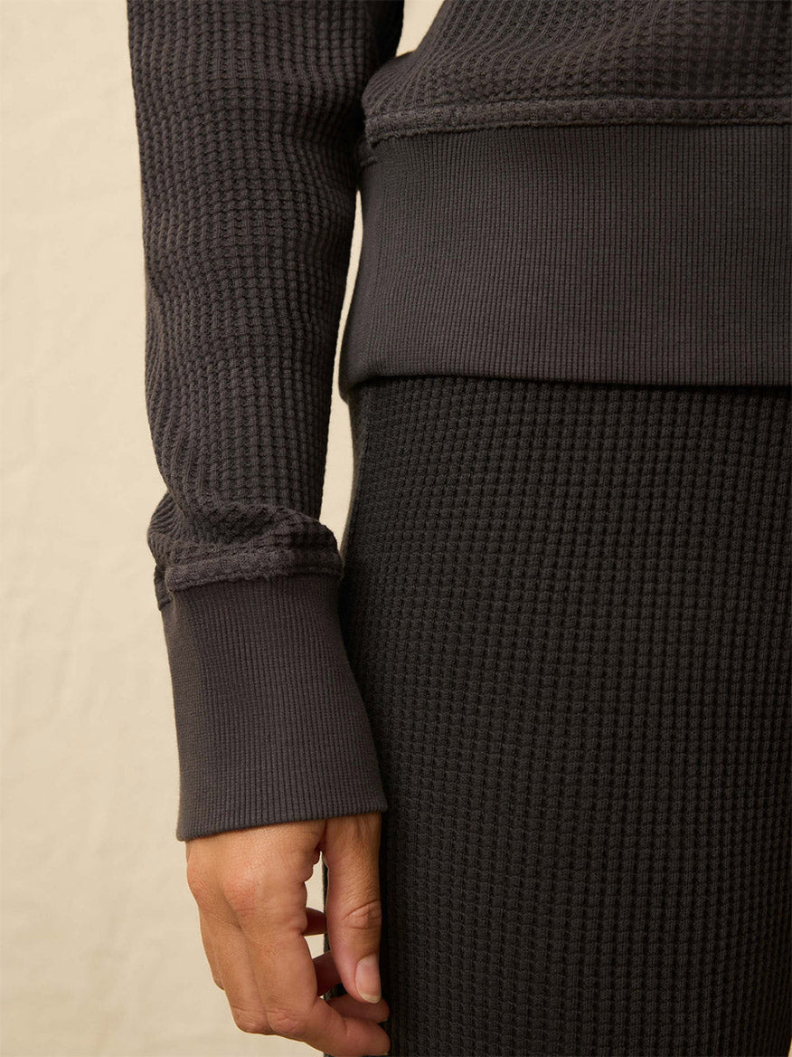 Close-up of a person’s hand resting by their side, wearing the Faherty Brand Après Waffle Henley in Washed Black with a matching skirt against a plain neutral background.