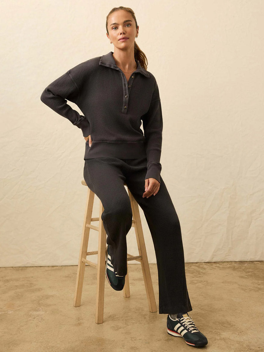Woman sits on a wooden stool, wearing the Faherty Brand Après Waffle Henley in Washed Black with matching pants and black sneakers against a neutral background.