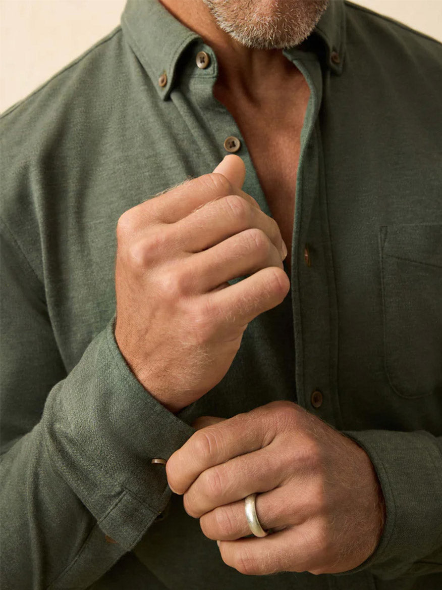 A man with a gray beard adjusts his cuff while wearing the Faherty Brand Coastline Knit Shirt in Forest Heather Twill. Only his hands, torso, and lower face show, emphasizing the button-down style and comfort of this casual shirt.