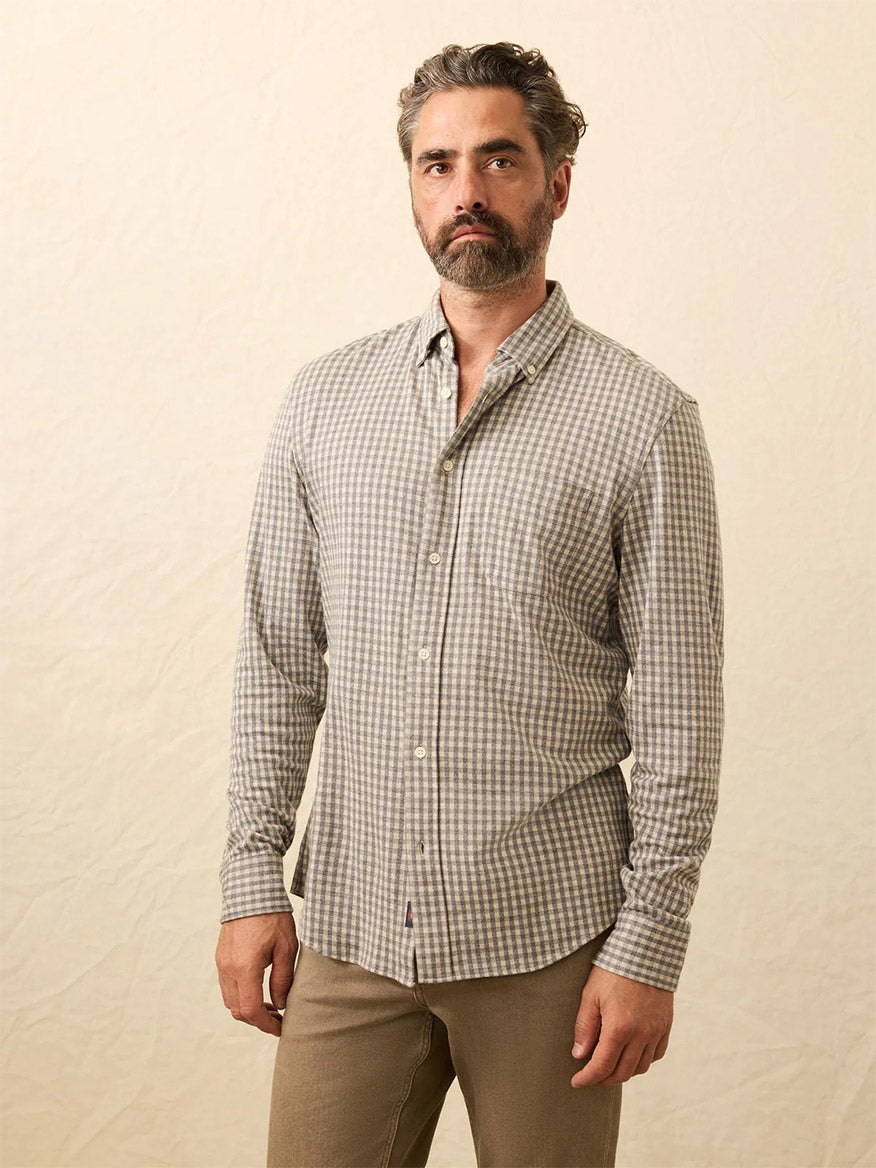 A man with gray hair and beard wears a Faherty Brand Coastline Knit Shirt in Fox Grey Gingham, paired with tan pants, as he stands against a plain light background.
