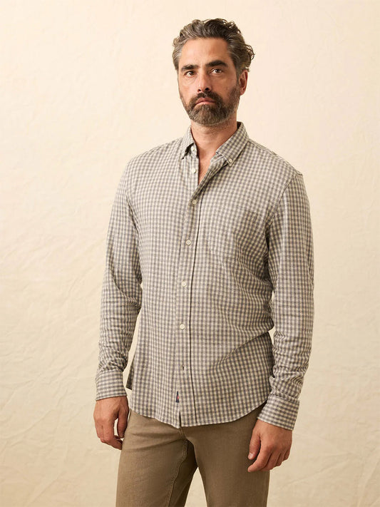 A man with gray hair and beard wears a Faherty Brand Coastline Knit Shirt in Fox Grey Gingham, paired with tan pants, as he stands against a plain light background.