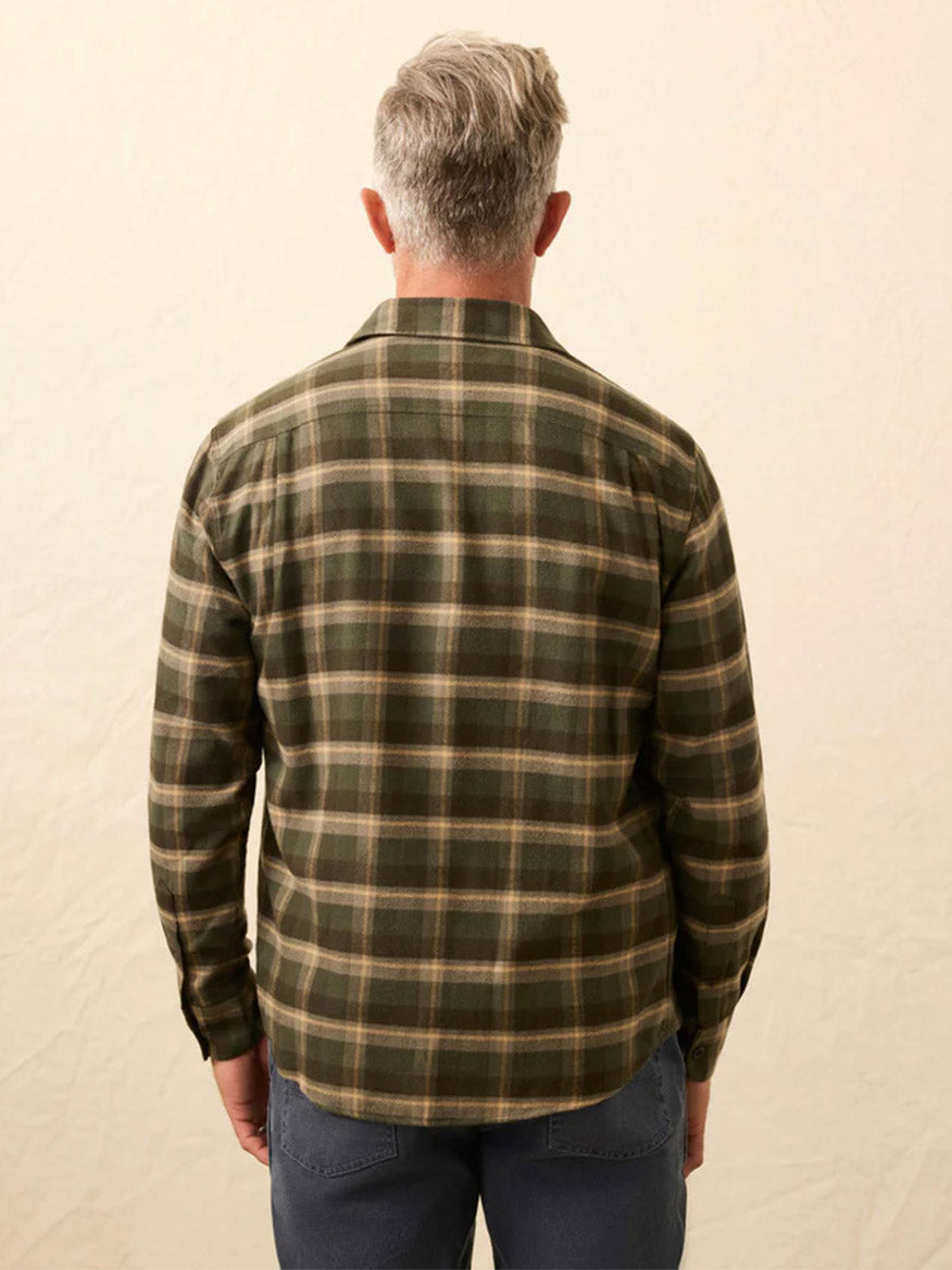 A person with gray hair stands with their back to the camera, wearing the Faherty Brand Heirloom Portuguese Flannel Shirt in Garrett Verde Plaid and dark pants against a plain, light background.
