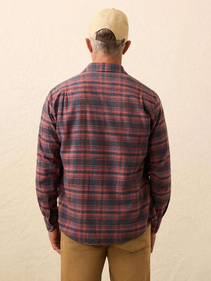 A man faces away against a plain light background, wearing a beige cap, classic fit tan pants, and the Faherty Brand Heirloom Portuguese Flannel Shirt in Porto Covo Plaid (red and blue).