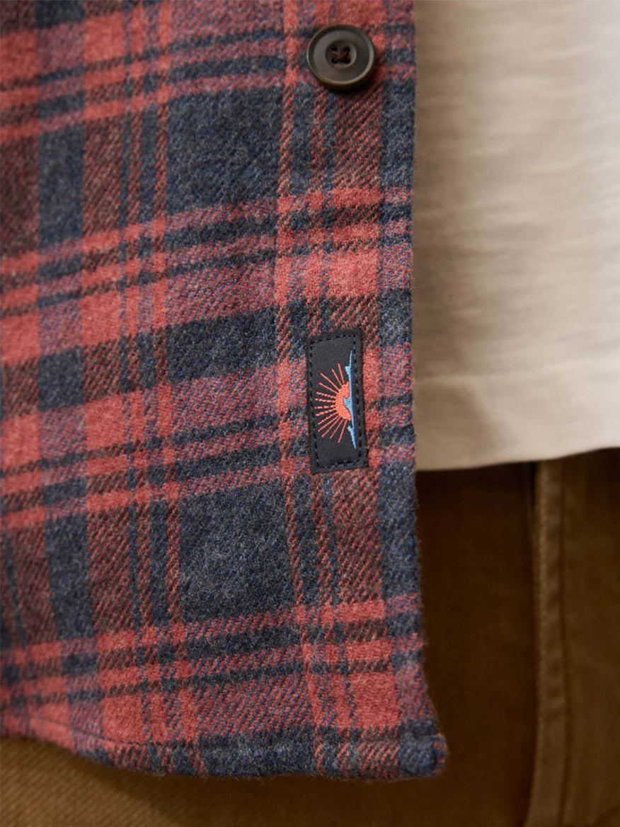 Close-up of the Faherty Brand Heirloom Portuguese Flannel Shirt in Porto Covo Plaid, featuring red and blue plaid, a button, and a sunburst tag, worn over a white shirt and brown pants; classic fit and Portuguese craftsmanship.