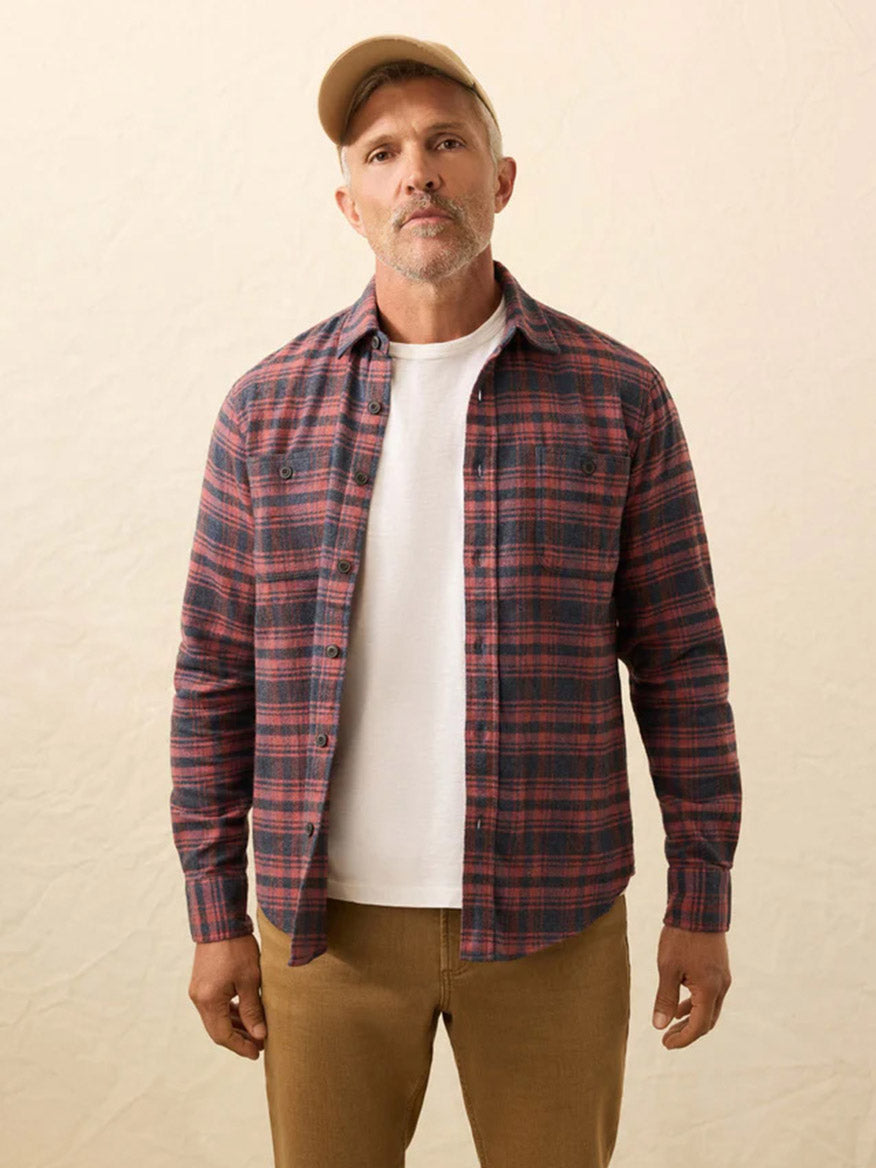 A man wearing a beige cap, tan pants, and a Faherty Brand Heirloom Portuguese Flannel Shirt in Porto Covo Plaid over a white t-shirt stands against a neutral background.